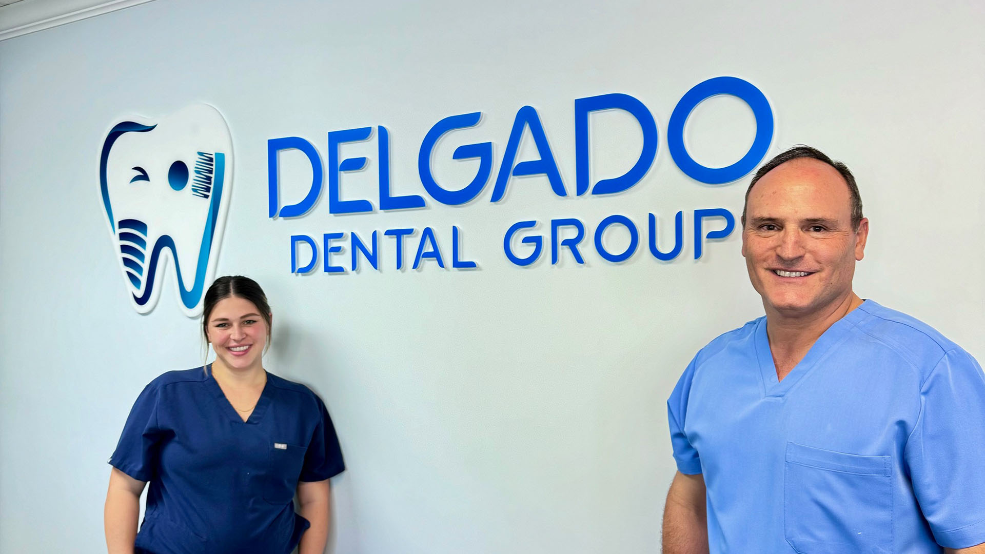 The image shows a professional setting with two individuals standing in front of a sign that reads  DELGADO DENTAL GROUP  and includes the logo of a dental company.