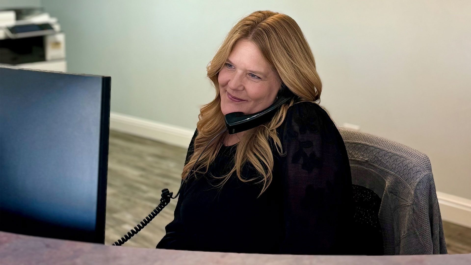 The image shows a woman sitting at a desk with a computer monitor, smiling and looking towards the camera. She is wearing a black top and has her hair styled in loose waves.