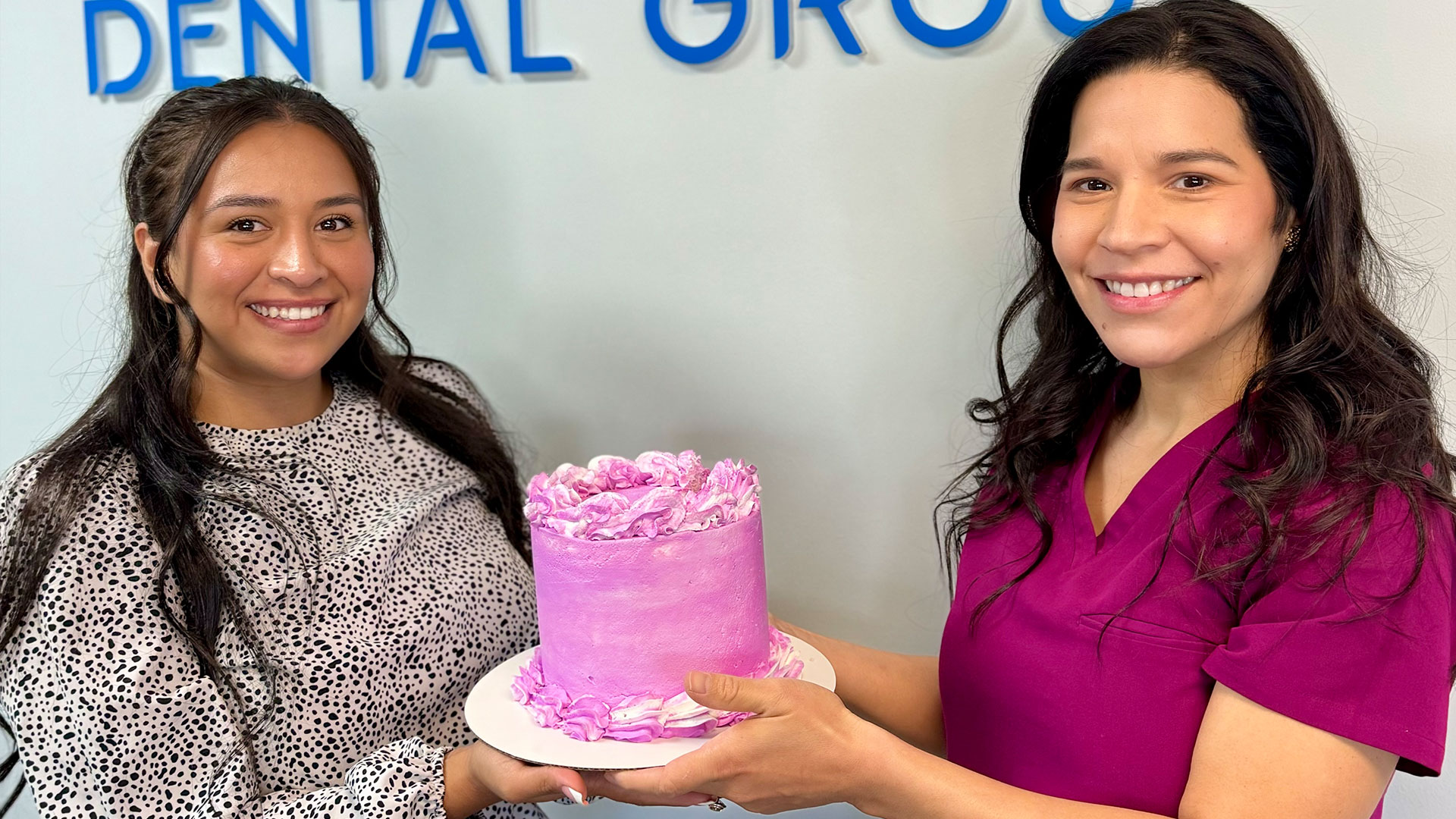 The image features two individuals, one holding a cake with pink frosting on a plate. They are standing in front of a sign that reads  DENTAL GROUP.