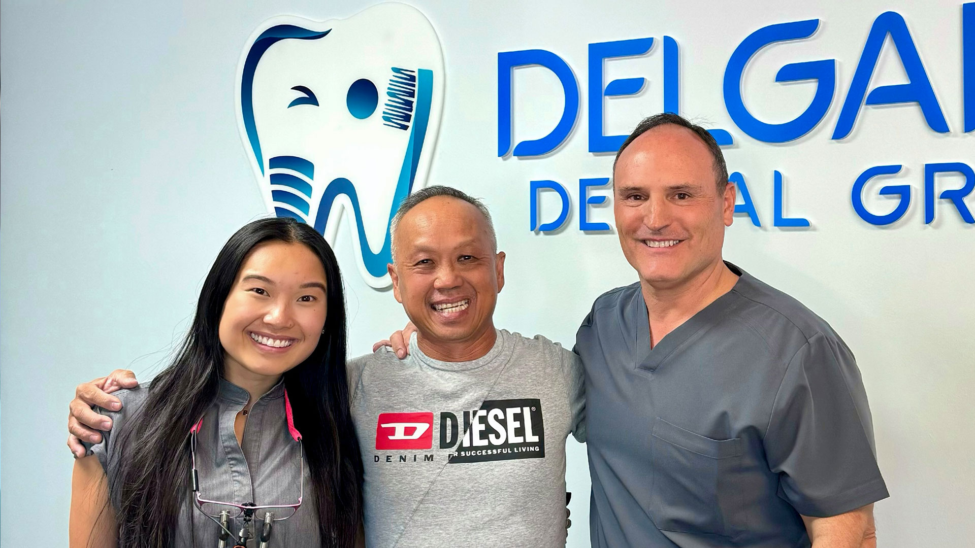 The image shows three individuals standing in front of a dental practice sign, smiling and posing for the photo.