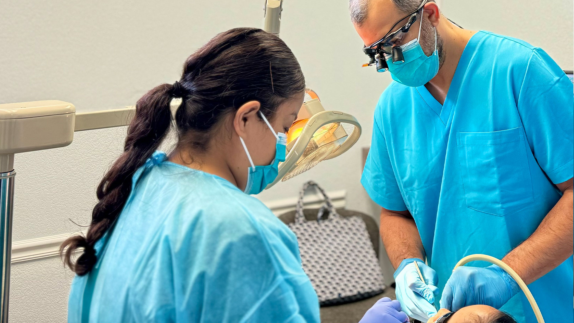 A man in a blue surgical gown and a woman in a white surgical gown are wearing face masks and are engaged in dental or medical surgery.