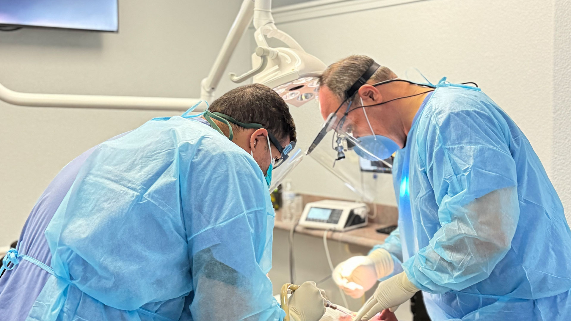 In the image, two individuals in medical scrubs and face masks are standing beside each other at a surgical table, with one person operating a device and the other observing.