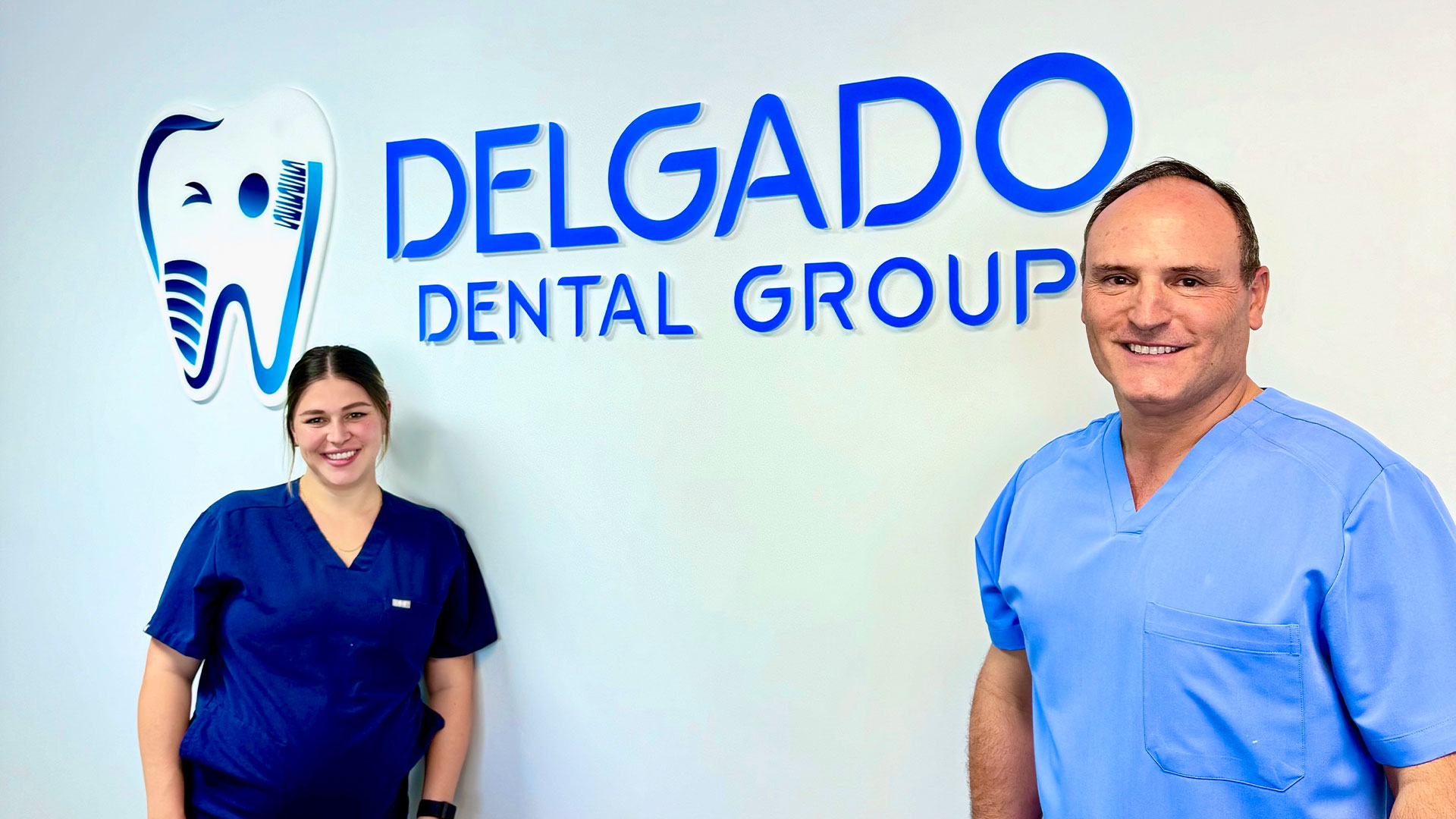 The image shows a man and a woman standing in front of a sign for  Delgado Dental Group.