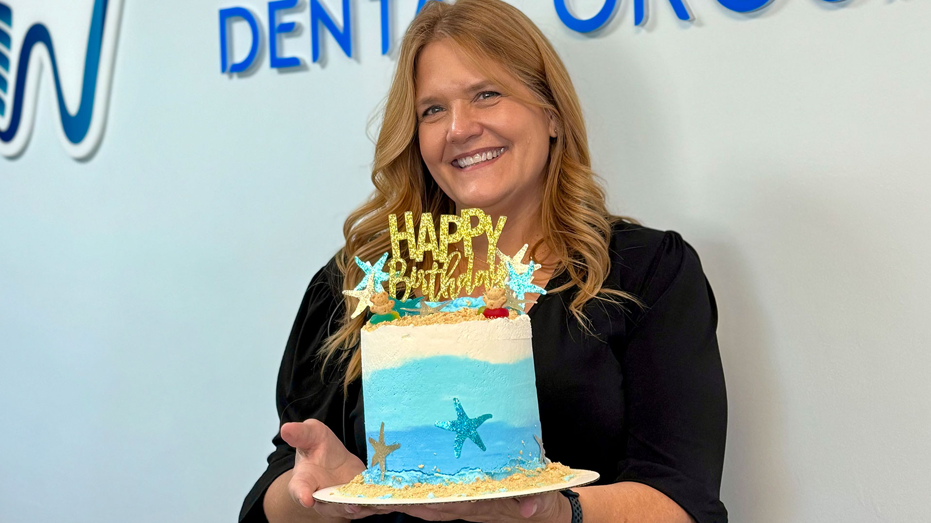 A woman is holding a cake with a  Happy Birthday  message and a beach theme, standing in front of a Dental Group sign.