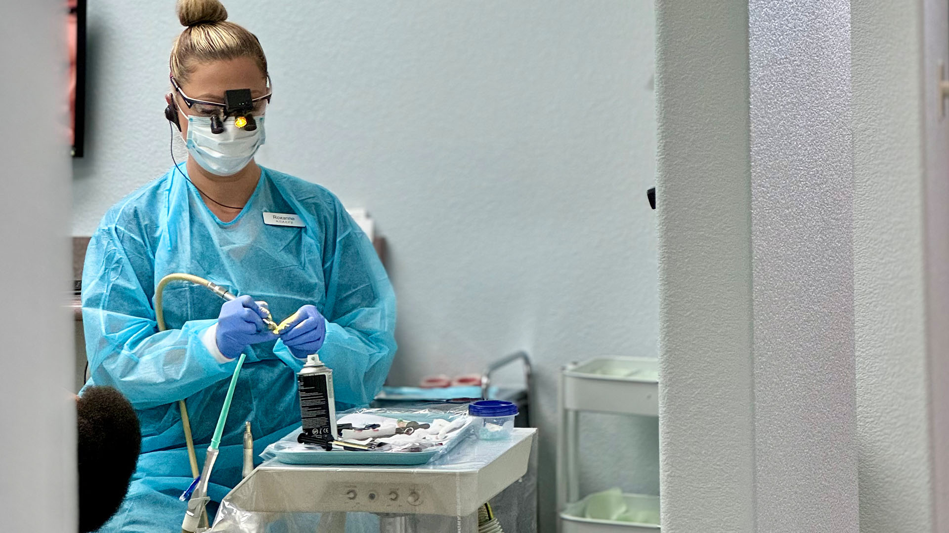 A person in a medical gown and mask, standing at a desk with various medical equipment.