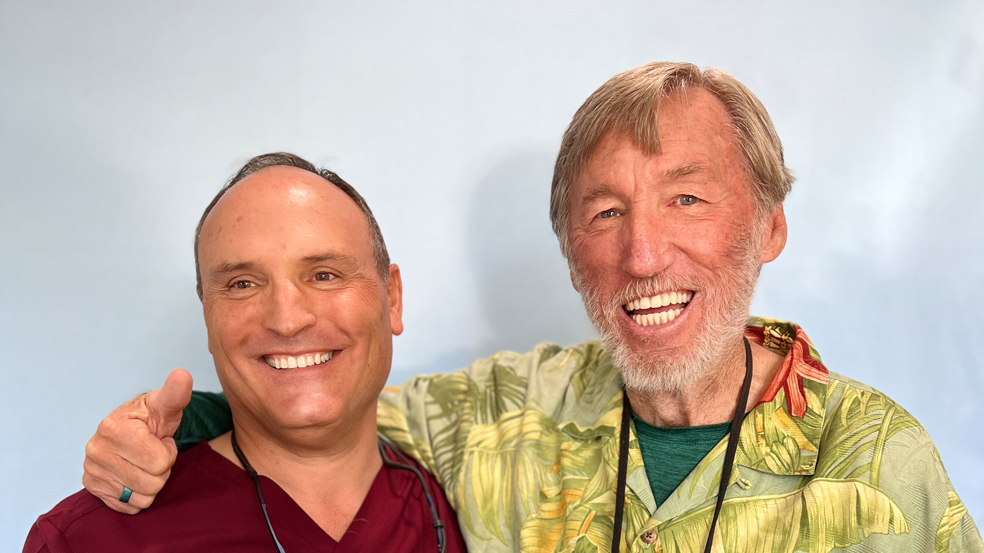 Two men posing for a photo, one with grey hair and wearing a Hawaiian shirt, the other with blond hair and a red tie.