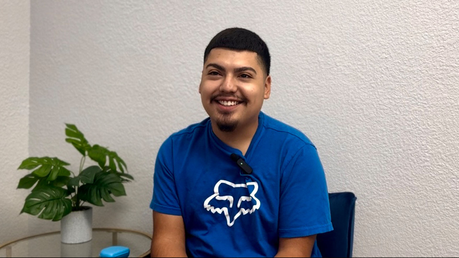 The image shows a man sitting in front of a plant, smiling at the camera.