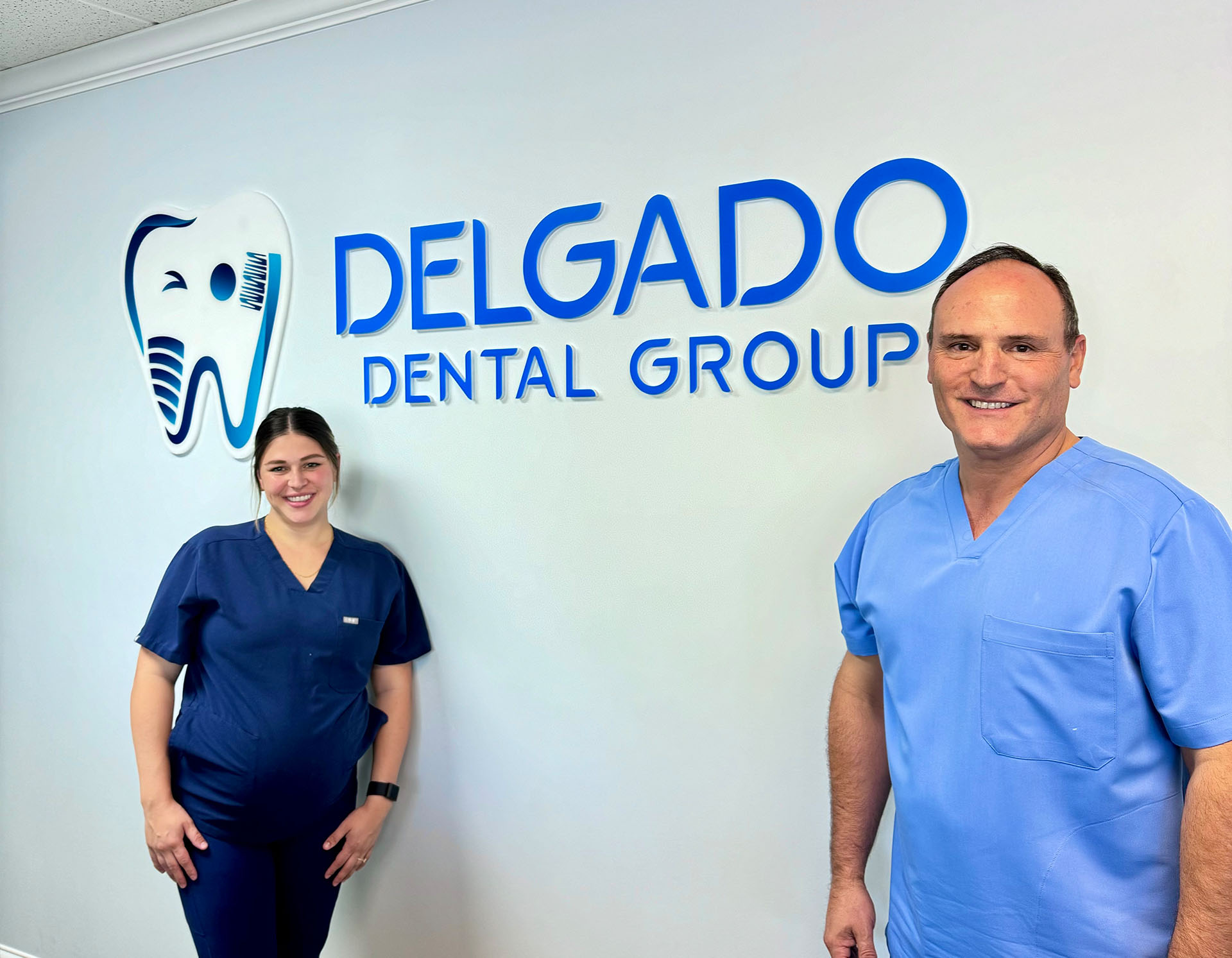 The image shows two individuals, a man and a woman, standing in front of a sign for  Delgado Dental Group.  They are posing for the photograph with smiles on their faces.